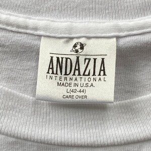 May include: White t-shirt tag with black text that reads "Andazia International Made in U.S.A. L (42-44) Care Over"