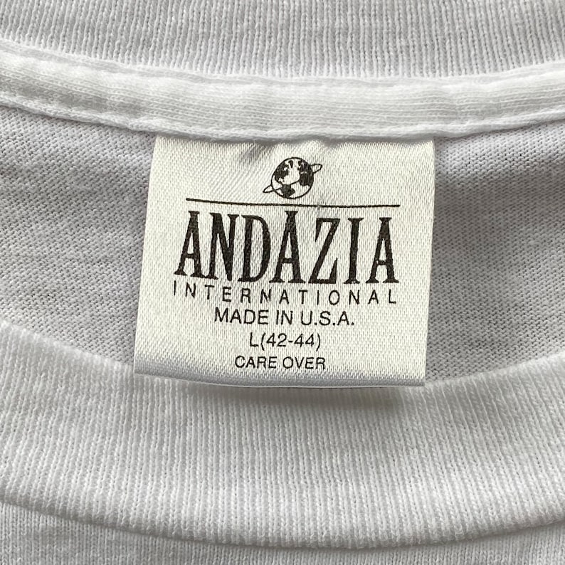 May include: White t-shirt tag with black text that reads "Andazia International Made in U.S.A. L (42-44) Care Over"