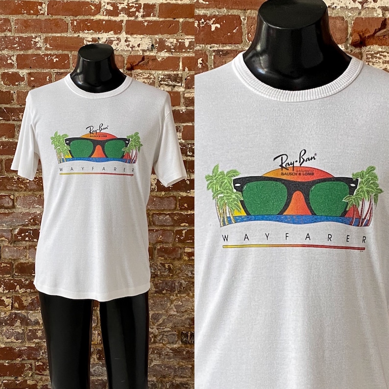 May include: White t-shirt with a graphic of Ray-Ban Wayfarer sunglasses with a sunset and palm trees. The text "Ray-Ban" and "Wayfarer" are printed on the shirt.
