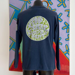 May include: Navy blue long-sleeve shirt with a circular graphic on the back. The graphic is green and white and features text that reads "Surf Curbs West Coast". The shirt is displayed on a black mannequin.