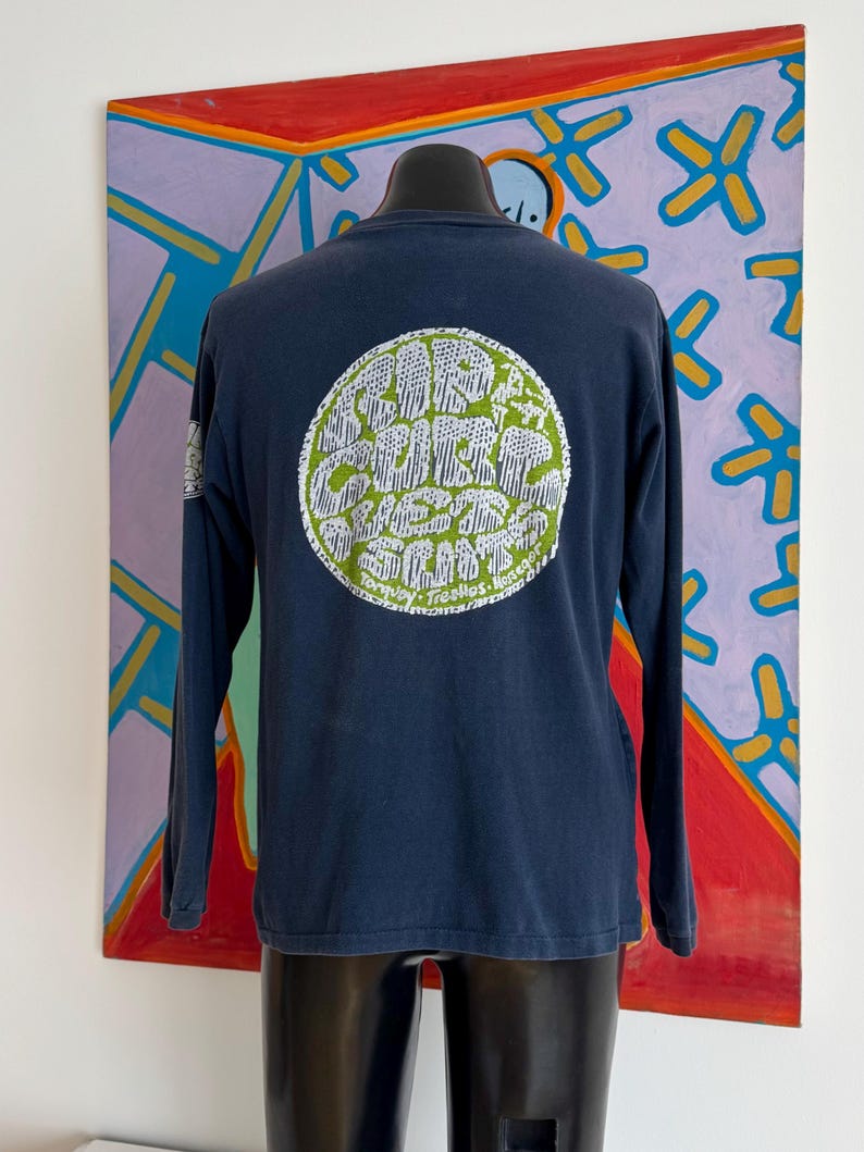 May include: Navy blue long-sleeve shirt with a circular graphic on the back. The graphic is green and white and features text that reads "Surf Curbs West Coast". The shirt is displayed on a black mannequin.