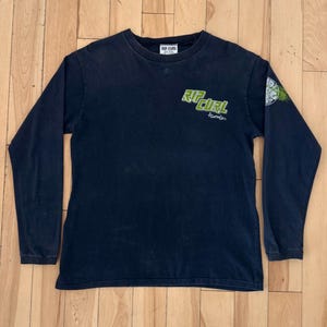 May include: Dark navy blue long-sleeve shirt with the text "RIP CURL" in green on the front and a small graphic on the sleeve. The shirt has a crew neck and a small tag at the neckline.