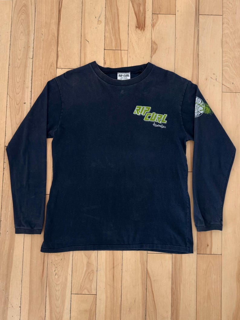 May include: Dark navy blue long-sleeve shirt with the text "RIP CURL" in green on the front and a small graphic on the sleeve. The shirt has a crew neck and a small tag at the neckline.