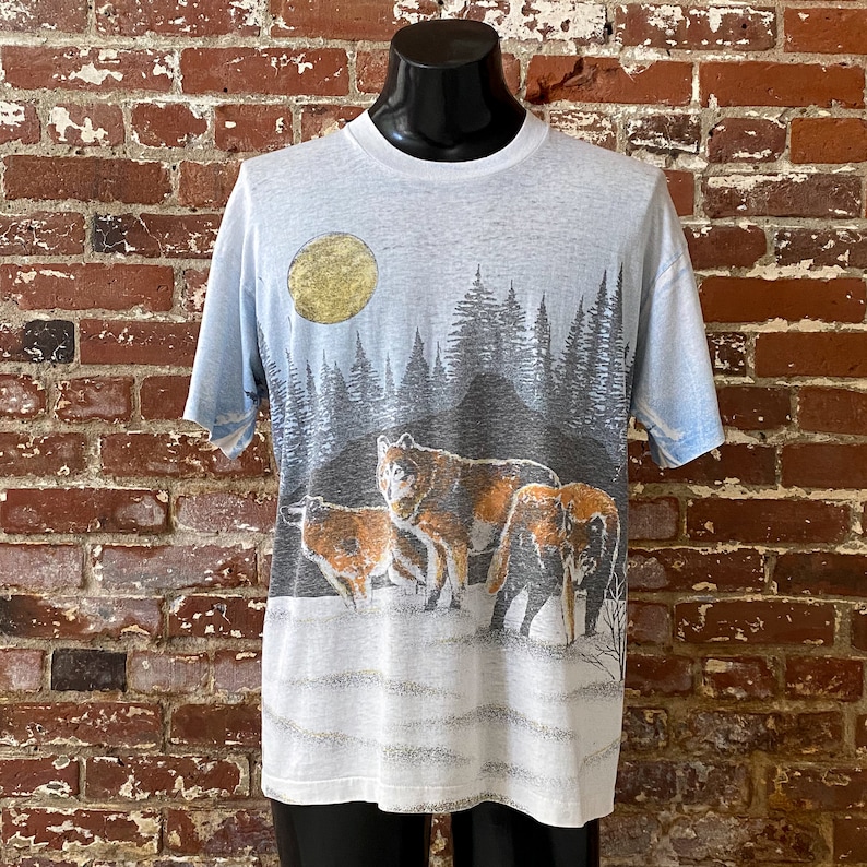 Puede incluir: Camiseta azul claro con un dise&ntilde;o de manada de lobos en una escena de bosque nevado bajo una luna dorada. El gr&aacute;fico incluye ilustraciones detalladas de lobos, &aacute;rboles y un cielo degradado. La camiseta se muestra en un maniqu&iacute; negro.