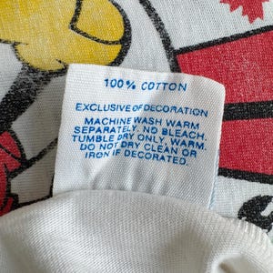 Może przedstawiać: Zbliżenie białej metki z tkaniny z niebieskim tekstem. Na metce widnieje napis "100% COTTON" i instrukcje prania. Tło przedstawia czerwoną i białą tkaninę we wzory z ż&oacute;łto-czarnym wzorem.