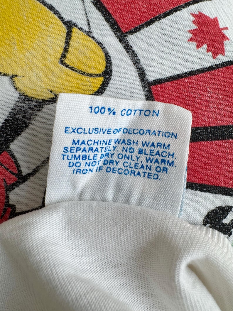 Może przedstawiać: Zbliżenie białej metki z tkaniny z niebieskim tekstem. Na metce widnieje napis "100% COTTON" i instrukcje prania. Tło przedstawia czerwoną i białą tkaninę we wzory z ż&oacute;łto-czarnym wzorem.