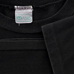 May include: Close-up of a black t-shirt with a white tag that says "Oasis" in green letters.