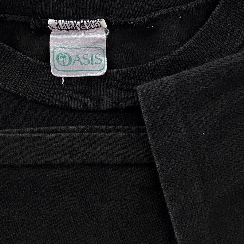 May include: Close-up of a black t-shirt with a white tag that says "Oasis" in green letters.