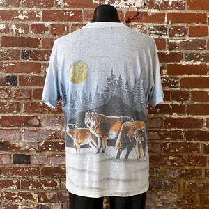 Puede incluir: Camiseta azul claro con una escena de manada de lobos. El dise&ntilde;o incluye una luna llena, un bosque y varios lobos en tonos marrones, grises y negros. La camiseta se muestra en un maniqu&iacute; contra una pared de ladrillos.