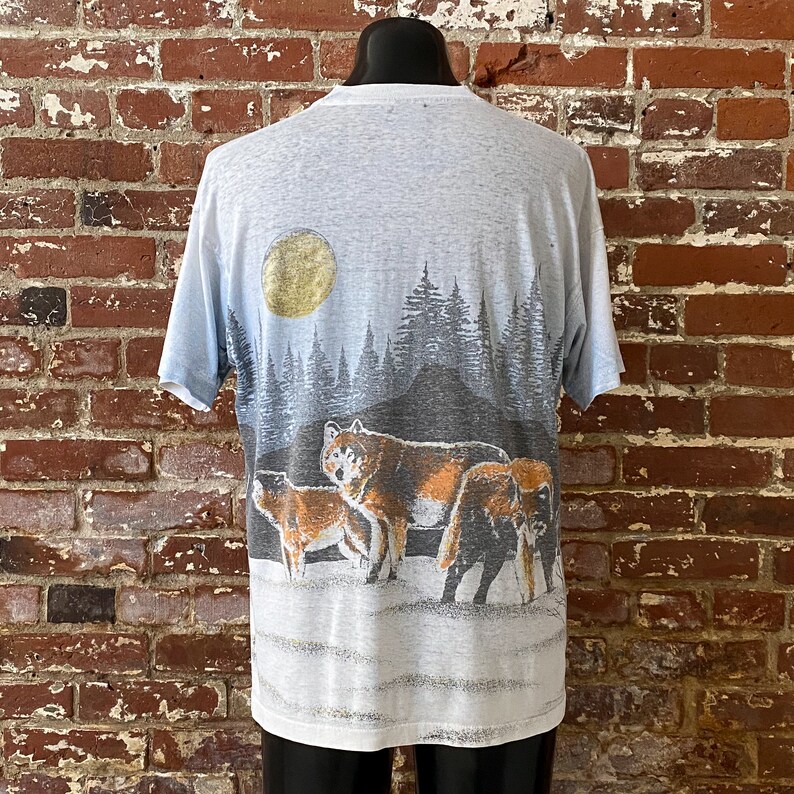 Puede incluir: Camiseta azul claro con una escena de manada de lobos. El dise&ntilde;o incluye una luna llena, un bosque y varios lobos en tonos marrones, grises y negros. La camiseta se muestra en un maniqu&iacute; contra una pared de ladrillos.