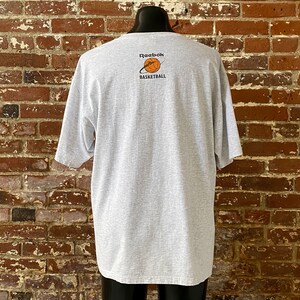 May include: A gray t-shirt with a Reebok Basketball logo on the back. The logo is a basketball with a white orbit around it. The text "Reebok Basketball" is below the basketball.