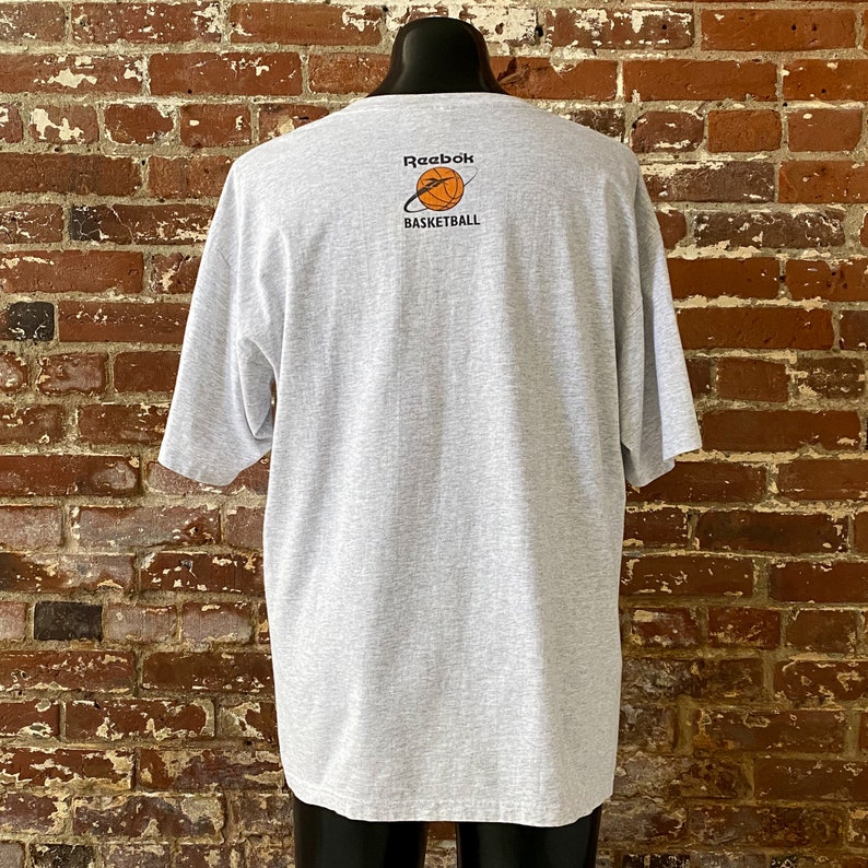 May include: A gray t-shirt with a Reebok Basketball logo on the back. The logo is a basketball with a white orbit around it. The text "Reebok Basketball" is below the basketball.