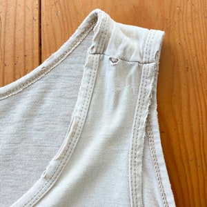 May include: Close-up of the shoulder seam of a light beige sleeveless top. The seam is frayed and has a small hole.