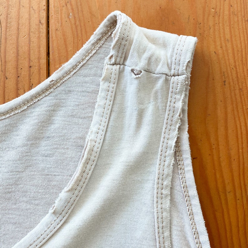 May include: Close-up of the shoulder seam of a light beige sleeveless top. The seam is frayed and has a small hole.