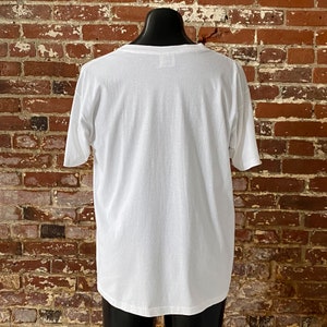 May include: A white t-shirt with a crew neck and short sleeves. The shirt is on a mannequin against a brick wall.