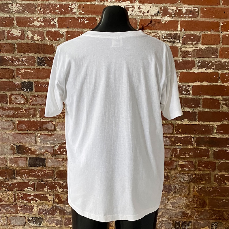 May include: A white t-shirt with a crew neck and short sleeves. The shirt is on a mannequin against a brick wall.