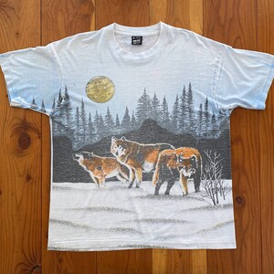 Puede incluir: Camiseta blanca con un dise&ntilde;o de paisaje que representa tres lobos en un campo nevado bajo una luna llena. El fondo muestra un bosque y un cielo degradado. La camiseta tiene cuello redondo y mangas cortas.