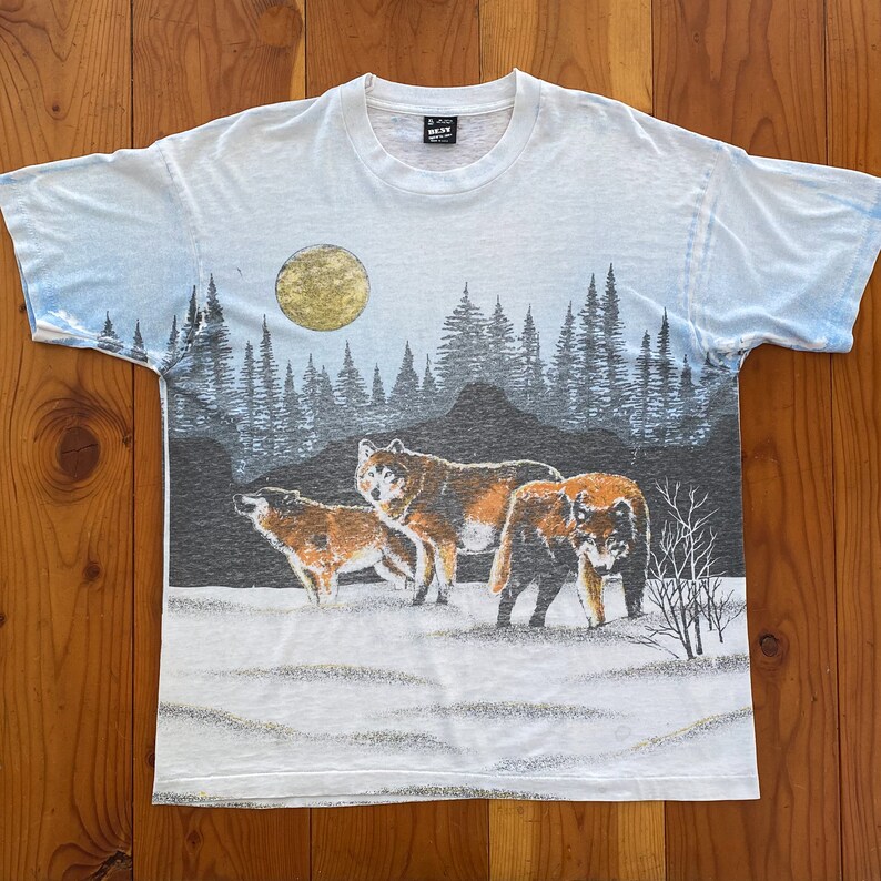 Puede incluir: Camiseta blanca con un dise&ntilde;o de paisaje que representa tres lobos en un campo nevado bajo una luna llena. El fondo muestra un bosque y un cielo degradado. La camiseta tiene cuello redondo y mangas cortas.