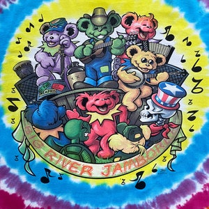 Op de afbeelding: Een tie-dye T-shirt met een kleurrijke illustratie van cartoonberen die muziekinstrumenten bespelen. De beren dragen hoeden en zonnebrillen. De tekst "Big River Jamboree" staat in een banner rond de beren. Het T-shirt is een levendige mix van blauw, geel, roze en groen.