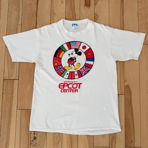 Może przedstawiać: Biała koszulka z Myszka Miki przed okręgiem z flagami międzynarodowymi. Tekst "EPCOT CENTER" jest wydrukowany na czerwono. Koszulka ma kr&oacute;tkie rękawy i okrągły dekolt.