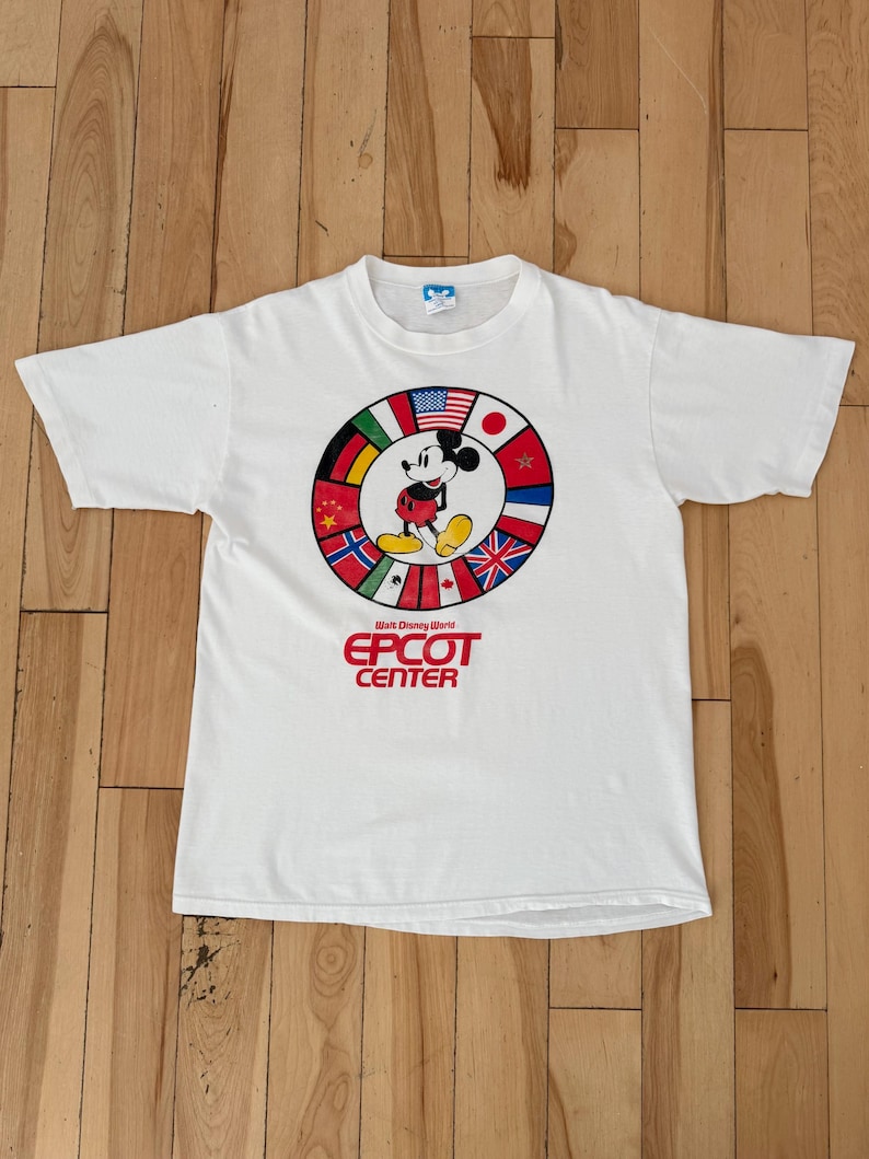 Może przedstawiać: Biała koszulka z Myszka Miki przed okręgiem z flagami międzynarodowymi. Tekst "EPCOT CENTER" jest wydrukowany na czerwono. Koszulka ma kr&oacute;tkie rękawy i okrągły dekolt.