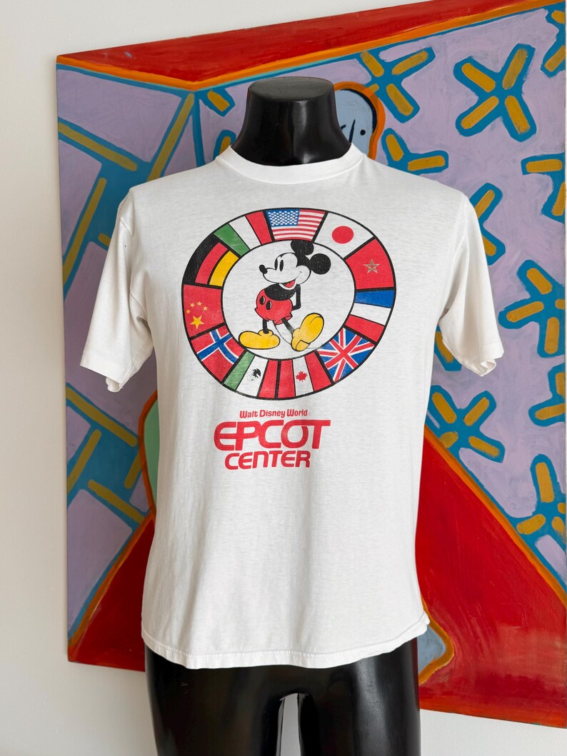 Może przedstawiać: Biała koszulka z Myszka Miki otoczonym flagami międzynarodowymi. Tekst "Walt Disney World" i "EPCOT CENTER" jest wydrukowany na czerwono. Koszulka jest prezentowana na czarnym manekinie.