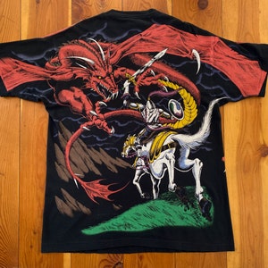 Może przedstawiać: Czarny t-shirt z fantastyczną sceną: czerwony smok atakuje rycerza na koniu. Rycerz trzyma miecz, a smok ma duże skrzydła. Projekt jest szczeg&oacute;łowy i kolorowy, na ciemnym tle.