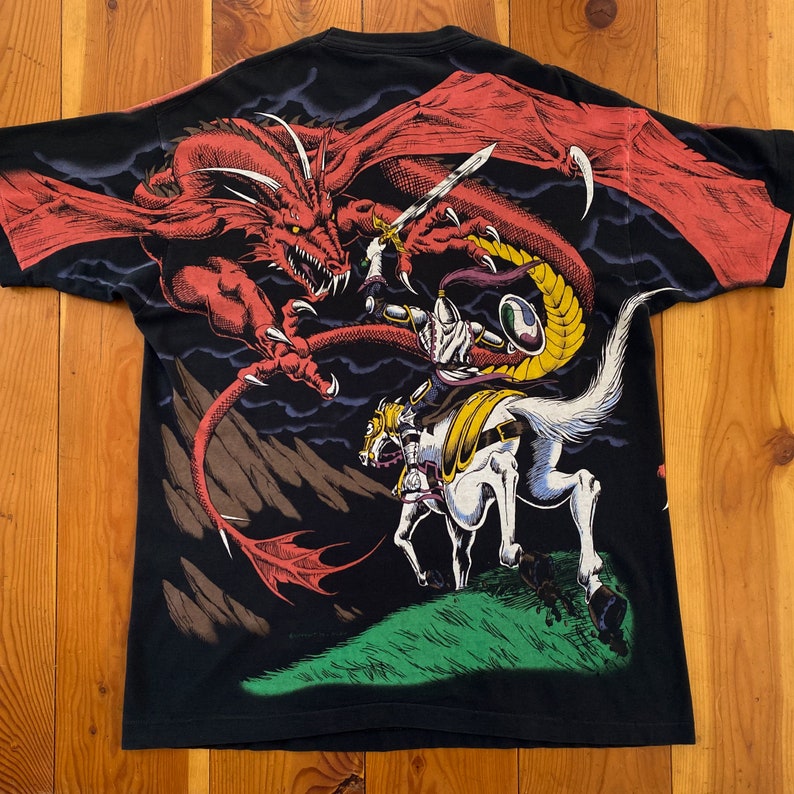 Może przedstawiać: Czarny t-shirt z fantastyczną sceną: czerwony smok atakuje rycerza na koniu. Rycerz trzyma miecz, a smok ma duże skrzydła. Projekt jest szczeg&oacute;łowy i kolorowy, na ciemnym tle.