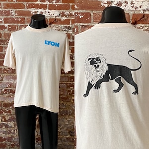 May include: Cream-colored t-shirt with the word "LYON" in blue block letters on the front. The back of the shirt features a black lion graphic. The shirt has a light tan collar and short sleeves. The shirt is displayed on a black mannequin.
