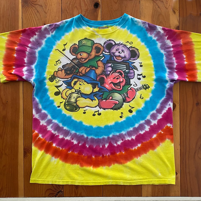 Op de afbeelding: Een tie-dye T-shirt met een cirkelvormig ontwerp met vier cartoonberen die muziekinstrumenten bespelen. De beren dragen hoeden en hebben verschillende kleuren vacht. Het shirt is geel, blauw, roze, oranje en paars.