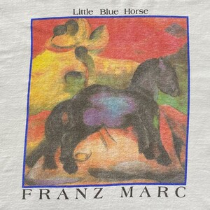 May include: A vintage t-shirt featuring a painting of a black horse in a colorful landscape. The text "Little Blue Horse" and "FRANZ MARC" are printed on the shirt.