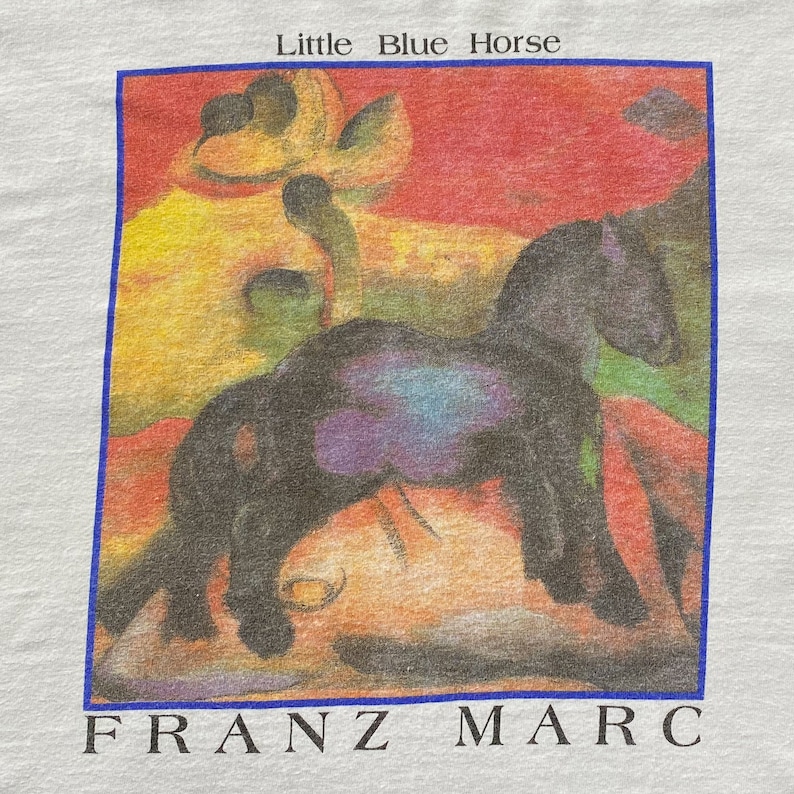 May include: A vintage t-shirt featuring a painting of a black horse in a colorful landscape. The text "Little Blue Horse" and "FRANZ MARC" are printed on the shirt.