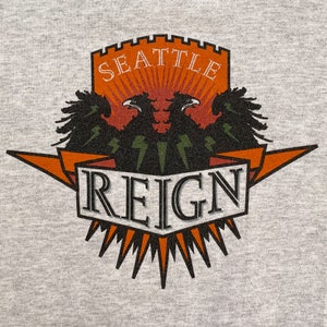 May include: A gray t-shirt with a graphic of a crest featuring two black eagles with orange and white accents. The crest reads "Seattle Reign" in a black and white font.