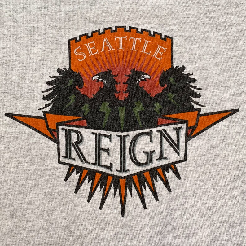 May include: A gray t-shirt with a graphic of a crest featuring two black eagles with orange and white accents. The crest reads "Seattle Reign" in a black and white font.