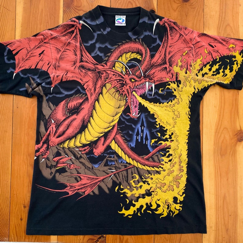 Może przedstawiać: Czarny t-shirt z czerwonym smokiem ziejącym ogniem. Smok ma ż&oacute;łte akcenty i jest osadzony na tle zamku i ciemnych chmur. Koszulka graficzna o tematyce fantasy.