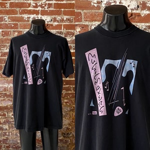 May include: Black t-shirt with a graphic of a bass guitar and the text "James Brown" in pink and blue. The text is stylized and appears to be written in a script font. The graphic is on the front of the shirt.