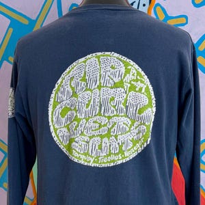 May include: Navy blue long-sleeve shirt with a large circular graphic on the back. The graphic features white text on a green background, with the words "Surf Club" and other text. The shirt is displayed on a mannequin.
