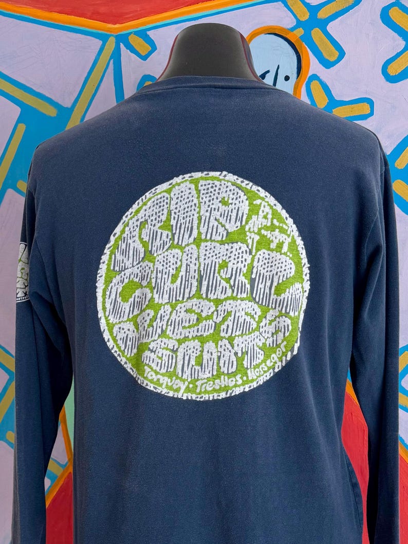 May include: Navy blue long-sleeve shirt with a large circular graphic on the back. The graphic features white text on a green background, with the words "Surf Club" and other text. The shirt is displayed on a mannequin.