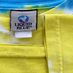 Op de afbeelding: Een geel, blauw en groen tie-dye T-shirt met een Liquid Blue-label. Het label zegt "Assembled in Mexico of U.S. Components" en "X-Large".