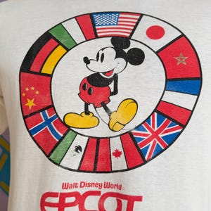 Może przedstawiać: Biała koszulka z Myszka Miki pośrodku okręgu z flagami międzynarodowymi. Na koszulce widnieje czerwony napis "Walt Disney World EPCOT CENTER". Myszka Miki ma na sobie czerwone spodenki, ż&oacute;łte buty i czarne rękawiczki.