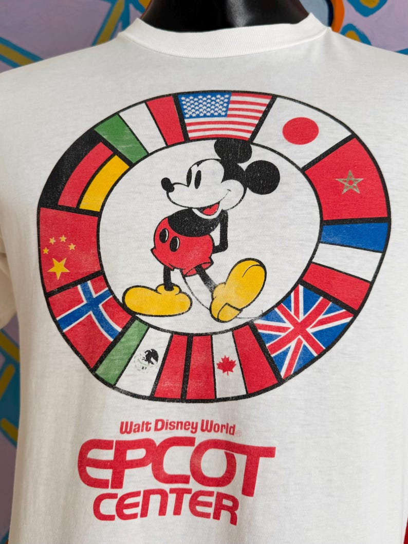Może przedstawiać: Biała koszulka z Myszka Miki pośrodku okręgu z flagami międzynarodowymi. Na koszulce widnieje czerwony napis "Walt Disney World EPCOT CENTER". Myszka Miki ma na sobie czerwone spodenki, ż&oacute;łte buty i czarne rękawiczki.