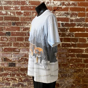 Puede incluir: Camiseta blanca con un dise&ntilde;o de paisaje invernal con lobos, &aacute;rboles y nieve. El gr&aacute;fico utiliza tonos grises, marrones y blancos, creando un dise&ntilde;o con tem&aacute;tica de la naturaleza. La camiseta se muestra en un maniqu&iacute; negro.