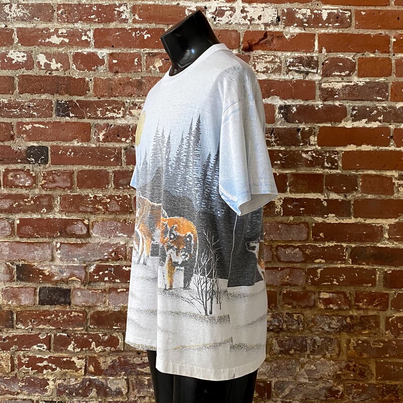 Puede incluir: Camiseta blanca con un dise&ntilde;o de paisaje invernal con lobos, &aacute;rboles y nieve. El gr&aacute;fico utiliza tonos grises, marrones y blancos, creando un dise&ntilde;o con tem&aacute;tica de la naturaleza. La camiseta se muestra en un maniqu&iacute; negro.