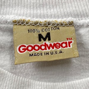 May include: A white tag with the text "100% Cotton" and "Goodwear" in red letters. The tag also has the letter "M" and the text "Made in U.S.A." in black letters.