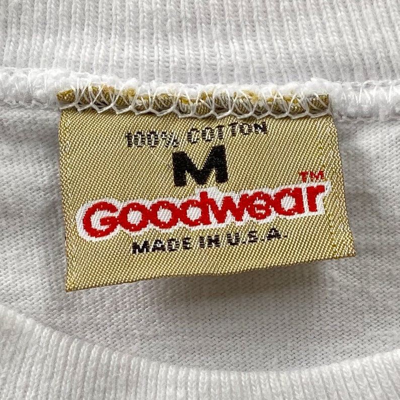 May include: A white tag with the text "100% Cotton" and "Goodwear" in red letters. The tag also has the letter "M" and the text "Made in U.S.A." in black letters.