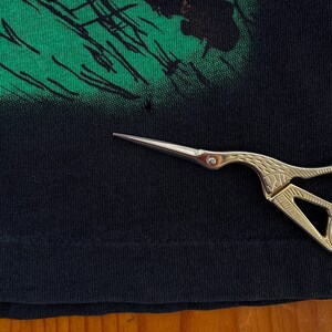 Może przedstawiać: Zbliżenie czarnej koszulki z zielonym wzorem graficznym. Złote nożyczki znajdują się na pierwszym planie, wskazując na małą dziurę w tkaninie. Koszulka leży na drewnianej powierzchni.