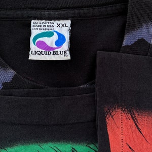 Może przedstawiać: Czarna koszulka Liquid Blue w rozmiarze XXL. Na metce napis "100% bawełna Made in USA Care on Reverse". Koszulka ma kolorowy wz&oacute;r graficzny z przodu i na rękawach. Logo Liquid Blue jest widoczne na metce.