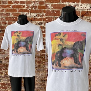 May include: White t-shirt with a colorful painting of horses and the text "Little Blue Horse" and "Franz Marc" printed on the front.