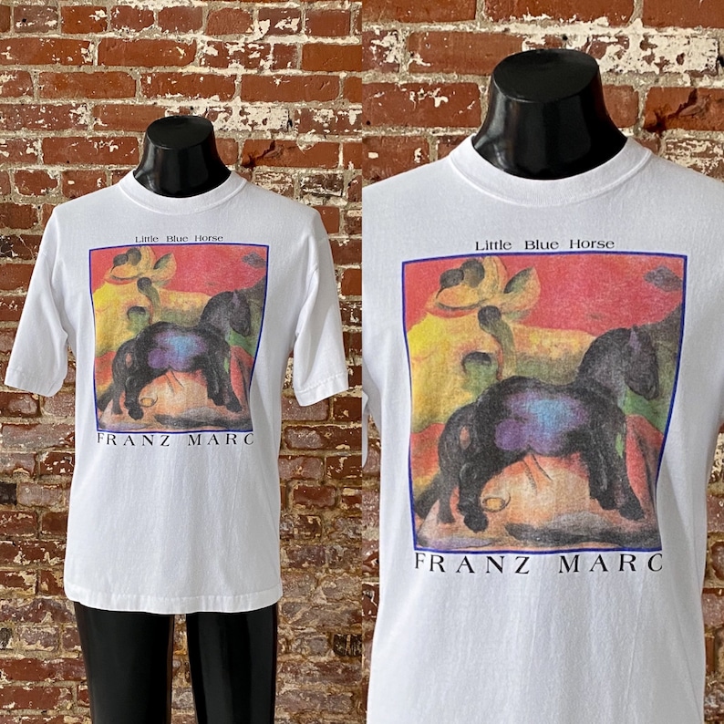 May include: White t-shirt with a colorful painting of horses and the text "Little Blue Horse" and "Franz Marc" printed on the front.