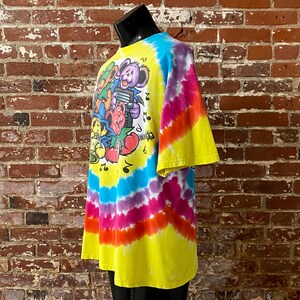 Op de afbeelding: Tie-dye T-shirt met een kleurrijke afbeelding van cartoonberen die muziekinstrumenten bespelen. Het shirt is geel, blauw, paars, oranje en rood.